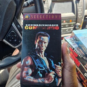 May include: A VHS tape of the movie Commando starring Arnold Schwarzenegger. The cover features a dark background with the actor in a black tactical vest holding a grenade, with the title Commando in yellow and red text at the top.