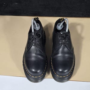 May include: A pair of black leather oxford shoes featuring a rounded toe, black laces, and a thick black sole with prominent yellow stitching along the welt. Each shoe includes a black heel pull tab with yellow text and a silver metal shoe tree insert.