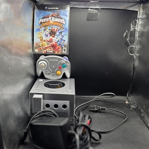 Può includere: Una console Nintendo GameCube color argento con controller argento abbinato, una custodia del gioco Power Rangers Dino Thunder e cavi di alimentazione e AV neri. La custodia presenta il titolo e illustrazioni dei personaggi.