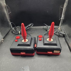 May include: Two black and red video game joysticks with a tall, ribbed red stick, two red buttons, and a black base. The text WICO COMMAND CONTROL is printed on the top, and one joystick features an AT-S200 label on the front red section.