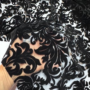 May include: Black velvet fabric with a floral damask pattern. The fabric is a sheer material with a raised, velvety design. The pattern is a repeating design of swirling leaves and flowers. The fabric is suitable for apparel and home decor.