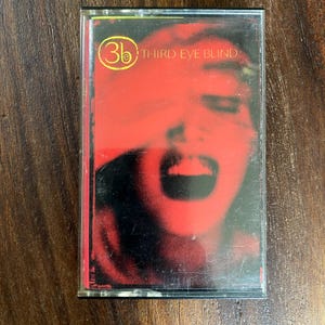 May include: A music cassette tape in a clear plastic case featuring the Third Eye Blind album cover. The artwork displays a high-contrast, red-toned close-up of a face with an open mouth and the yellow 3eb logo in the upper left corner.