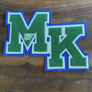May include: Two embroidered patches featuring the letters "M" and "K" in green with white trim, set against a blue background. The "M" patch has a small emblem in the center. These patches are likely for apparel or crafts.