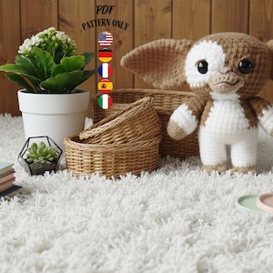 Op de afbeelding: Een gehaakt Gremlin-achtig wezen met grote oren, bruine en witte kleuren en grote ogen. De afbeelding bevat ook een witte potplant, een stapel boeken, gevlochten manden en kleine decoratieve dienbladen. De tekst "PDF PATTERN ONLY" is zichtbaar.