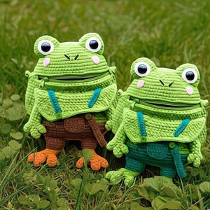 May include: Two handmade crochet frog-shaped bags. The bags are primarily green with large, round eyes, pink cheeks, and a zippered mouth. They have brown or teal shorts with orange or green feet, and blue suspenders. The bags are sitting in green grass.