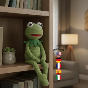 Puede incluir: Una muñeca de rana verde tejida a ganchillo con boca roja y ojos blancos está sentada en un estante de madera. La rana está al lado de una pequeña suculenta en maceta y una fila de libros. El estante es parte de una estantería.