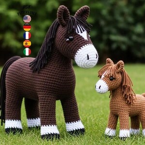 May include: Two crocheted horse figures, a large dark brown one and a smaller light brown one, stand on green grass. The larger horse has a white face and legs, with a black mane and tail. The smaller horse has a white face and legs, with a light brown mane and tail.