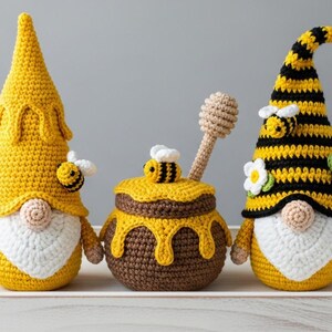 May include: Three handmade crochet bee-themed gnomes and a honey pot on a white tray. The gnomes have yellow hats, white beards, and bee accents. The honey pot is brown with yellow honey dripping down. A crochet honey dipper rests inside.