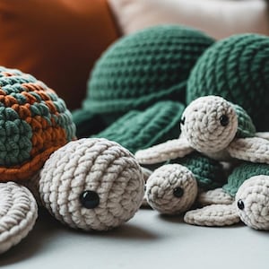 May include: A collection of crocheted sea turtles in various sizes. One large turtle features a shell with alternating sage green and burnt orange yarn. Several smaller turtles have cream-colored heads and flippers with dark green shells.