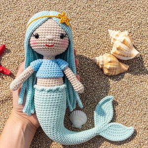 May include: A handmade crochet mermaid doll with blue hair, a blue top, and a light blue tail. A red starfish and seashells are next to the doll on a sandy surface. The doll has a gold star on its head.