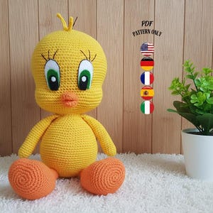 Può includere: Una bambola di Tweety Bird all'uncinetto gialla fatta a mano con piedi arancioni. La bambola ha grandi occhi verdi, lunghe ciglia e un becco arancione. Una piccola pianta in vaso si trova a destra della bambola. Il testo "PDF PATTERN ONLY" è visibile.