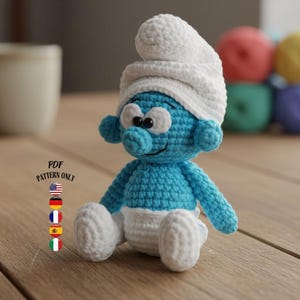 Può includere: Bambola dei Puffi all'uncinetto, con corpo blu, cappello bianco e pannolino bianco. La bambola ha occhi neri e un'espressione sorridente. L'immagine include anche una tazza da caffè e gomitoli di lana colorati. Il testo "PDF PATTERN ONLY" è visibile.