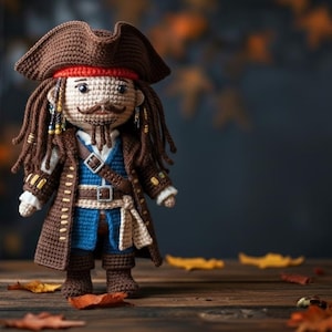Pode incluir: Boneca de crochê de um personagem pirata, com um chapéu de três pontas marrom, cabelo castanho comprido com contas e um bigode. A boneca usa um casaco marrom, um colete azul e um cinto. O fundo inclui folhas de outono.