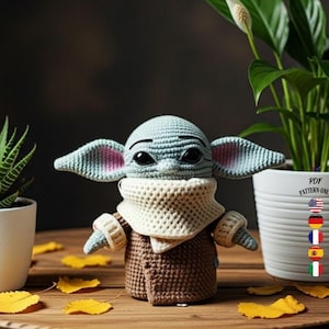 Op de afbeelding: Een gehaakte figuur van een personage met grote oren en grote ogen, gekleed in een bruine jas en een crèmekleurige sjaal. De figuur staat op een houten tafel met twee potplanten en gele bladeren.