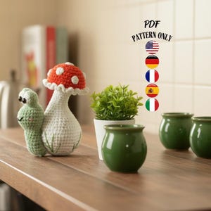 Puede incluir: Una figura de ganchillo de un caracol y una seta con un sombrero rojo y blanco. El caracol es verde con ojos negros. Junto a ellos hay pequeñas macetas de cerámica verde y una pequeña planta en maceta. El texto "PDF PATTERN ONLY" es visible.