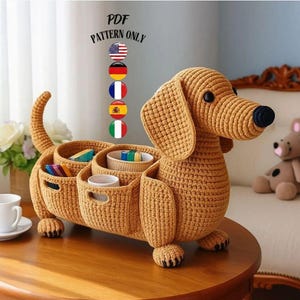 Peut inclure: Un panier de rangement en forme de teckel crocheté, de couleur beige. Le chien a un long corps avec plusieurs compartiments pour organiser des objets. Le texte "PDF PATTERN ONLY" est visible. Le nez du chien est noir.