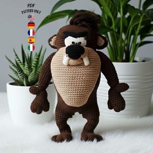 Pode incluir: Uma figura de Taz de crochê feita à mão em tons de marrom, bege, preto e branco. O personagem de desenho animado tem uma boca grande bege, dentes brancos e um nariz preto. A figura está em frente a plantas em vasos.