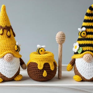 May include: Crocheted bee-themed gnomes, honey pot, and honey dipper. The gnomes have yellow and brown hats, white beards, and bee accents. The honey pot is brown with yellow honey dripping down. The honey dipper is light brown.