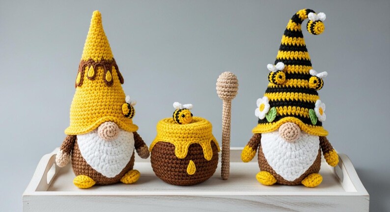 May include: Crocheted bee-themed gnomes, honey pot, and honey dipper. The gnomes have yellow and brown hats, white beards, and bee accents. The honey pot is brown with yellow honey dripping down. The honey dipper is light brown.