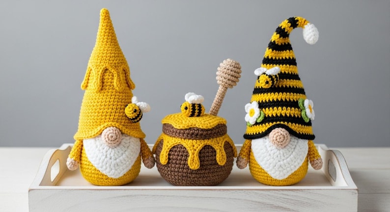 May include: Three handmade crochet bee-themed gnomes and a honey pot on a white tray. The gnomes have yellow and black striped hats, white beards, and bee accents. The honey pot is brown with yellow honey dripping down the sides. A crochet honey dipper rests inside.