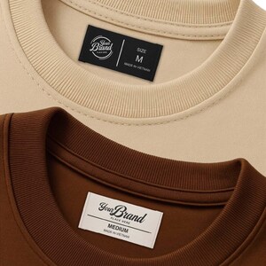 May include: Close-up of two crew neck sweatshirts, one beige and one brown. Each sweatshirt has a tag with the brand logo and size information. The beige sweatshirt tag reads "SIZE M". The brown sweatshirt tag reads "MEDIUM". Both tags say "MADE IN VIETNAM".