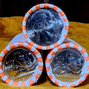 May include: Three rolls of U.S. quarters, each wrapped in orange and white striped paper. The top roll displays a portrait of George Washington. The bottom two rolls feature Anna May Wong. The coins are stacked on a gold-colored surface.