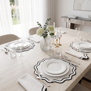 Può includere: Una tavola apparecchiata con piatti bianchi impilati su tovagliette rotonde con bordo nero smerlato. Ogni posto tavola include un tovagliolo bianco con bordo nero smerlato, posate in argento e bicchieri trasparenti. Un vaso centrale contiene fiori bianchi.
