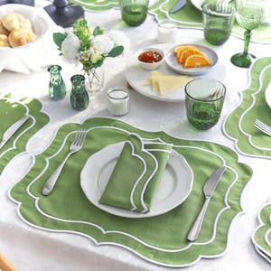 Könnte beinhalten: Ein gedeckter Tisch mit grünen Tischsets, Servietten und Gläsern. Weiße Teller, Besteck und ein kleiner Strauß weißer Blumen sind ebenfalls vorhanden. Der Tisch ist für eine Mahlzeit mit Orangen und Käse gedeckt.