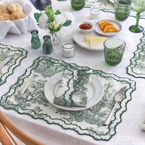 Può includere: Una tavola apparecchiata con una tovaglia bianca e dettagli verdi. Include un piatto bianco con un tovagliolo piegato, un sottopiatto con un disegno di albero verde, bicchieri verdi e piccoli piatti di cibo. Sono presenti anche un cestino di pane e un vaso di fiori.