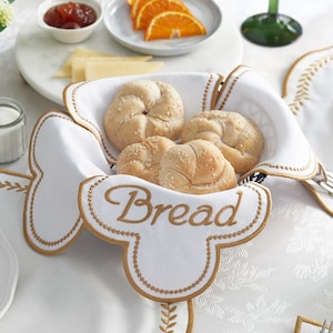 Könnte beinhalten: Ein weißer Brotkorb mit goldener Stickerei, gefüllt mit Brötchen. Das Wort "Bread" ist goldfarben gestickt. Der Korb steht auf einer weißen Tischdecke mit floralem Muster. Orangenstücke, Käse und Marmelade befinden sich auf einem Serviertablett aus Marmor.