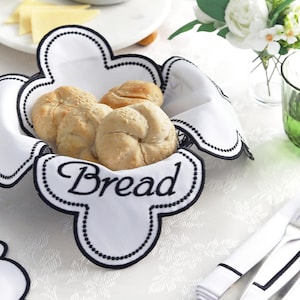 May include: A white bread basket with a black scalloped trim and the word "Bread" in script. The basket contains several golden-brown rolls. The scene includes a white tablecloth, silverware, and a small bouquet of white flowers.
