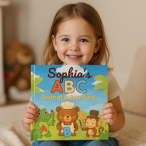 May include: A young girl smiles while holding a children's book titled "Sophia's ABC Animal Adventure." The book cover features colorful illustrations of animals, including a crocodile, bear, and monkey, with the letters A, B, and C.