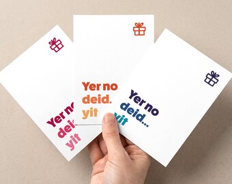 Puede incluir: Tres tarjetas de felicitación blancas con el texto "Yer no deid. Yit" en varios colores. Cada tarjeta tiene un pequeño icono de caja de regalo en un color a juego. Las tarjetas están sujetas por una mano.