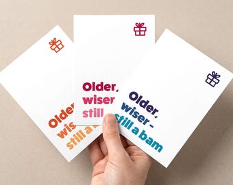 Puede incluir: Tres tarjetas de felicitación blancas con el texto "Older, wiser, still a bam" en varios colores. Cada tarjeta tiene un pequeño icono de caja de regalo en la esquina. Las tarjetas son sostenidas por una mano.