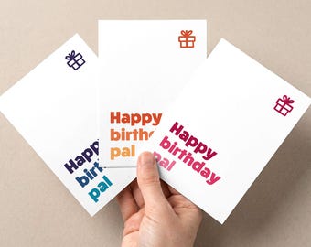 Puede incluir: Tres tarjetas de felicitación blancas con el texto "Happy birthday pal" en diferentes colores. Cada tarjeta tiene un pequeño icono de caja de regalo. Las tarjetas son sostenidas por una mano.