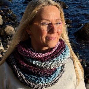 May include: A multi-colored knitted cowl in shades of purple, blue, and gray. The cowl is worn around the neck and is made of a soft, textured yarn. The background shows a person with blonde hair and glasses, with a body of water and rocks in the background.