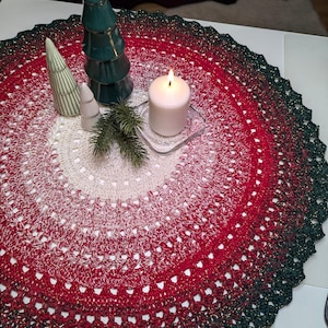 Può includere: Una tovaglia rotonda all'uncinetto con una sfumatura di colori, che passa dal crema al rosso e termina con un bordo verde scuro. Alberi di Natale in ceramica decorativi e una candela accesa sono posti sulla tovaglia, creando una scena festosa.