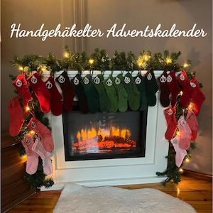 Op de afbeelding: Een witte open haard versierd met een slinger van groen en lichtslingers, versierd met een verzameling rode, groene en roze gebreide adventskalenderkousen, elk genummerd. De tekst "Handgehäkelter Adventskalender" staat bovenaan.