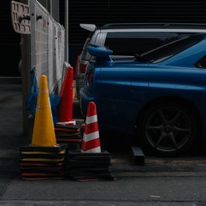 May include: A blue sports car is parked next to traffic cones in a parking area. The cones are yellow, red, and red and white striped. The image also includes a white fence and a silver SUV.