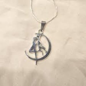 May include: A silver pendant necklace with a figure silhouette within a crescent moon. The pendant hangs from a delicate silver chain. The design is intricate, with the figure appearing to be dancing or in motion. The necklace is a delicate piece of jewellery.