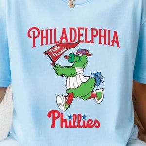 Może przedstawiać: Jasnoniebieska koszulka z napisem "PHILADELPHIA" w kolorze czerwonym, nad kresk&oacute;wkową maskotką trzymającą flagę z napisem "Phillie Phanatic". Słowo "Phillies" znajduje się pod maskotką w kolorze czerwonym.
