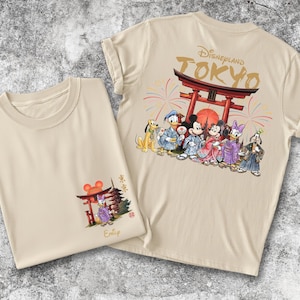 May include: Two beige t-shirts with Japanese-themed Disney graphics. One shirt features a front graphic of a red torii gate and a Disney character. The other shirt has a back graphic of Disney characters in traditional Japanese attire, with the text "Disneyland TOKYO".