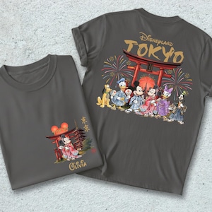 May include: Two gray t-shirts with Japanese-themed Disney designs. One shirt features Mickey and Minnie Mouse in traditional Japanese attire, with the name "Olivia" below. The other shirt has a larger design of Disney characters in front of a Torii gate with fireworks.
