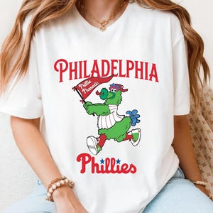 Może przedstawiać: Biała koszulka z logo Philadelphia Phillies w kolorze czerwonym i maskotką drużyny, Phillie Phanatic, w kolorze zielonym, niebieskim i białym. Napis "PHILADELPHIA" i "Phillies" jest czerwony, a maskotka trzyma flagę.