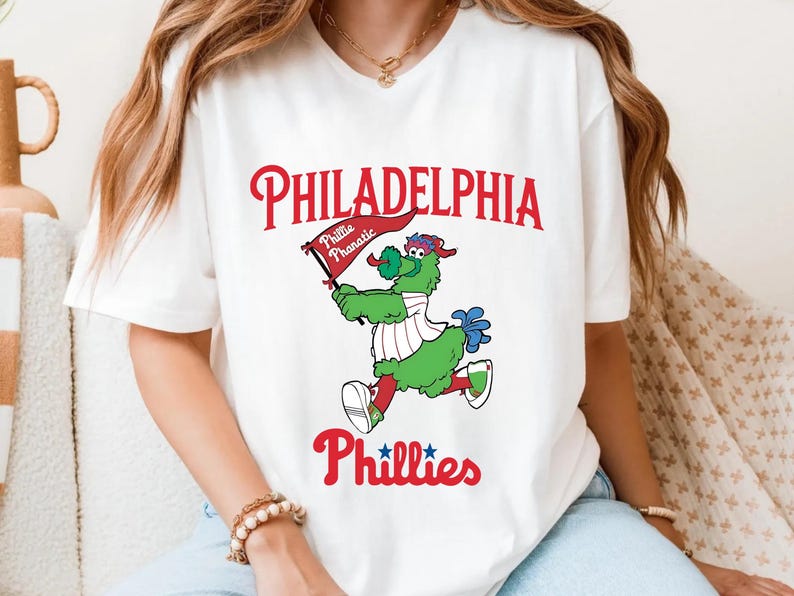 Może przedstawiać: Biała koszulka z logo Philadelphia Phillies w kolorze czerwonym i maskotką drużyny, Phillie Phanatic, w kolorze zielonym, niebieskim i białym. Napis "PHILADELPHIA" i "Phillies" jest czerwony, a maskotka trzyma flagę.