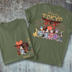 May include: Two olive green t-shirts. One features Mickey Mouse in a kimono in front of a Japanese pagoda. The other has a Disneyland Tokyo design with Disney characters in kimonos, fireworks, and a red torii gate.