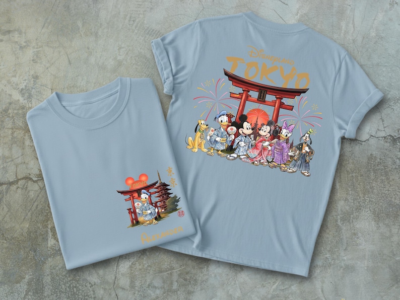 May include: Two light blue t-shirts with graphics. One shirt features a graphic of Mickey Mouse, Donald Duck, and other Disney characters in traditional Japanese attire in front of a red torii gate. The other shirt has a similar design.