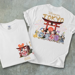 May include: Two white t-shirts with colorful graphics. One shirt features Mickey and Minnie Mouse in traditional Japanese attire in front of a red torii gate. The other shirt has the Disneyland Tokyo logo and various Disney characters in Japanese clothing.