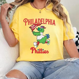 Może przedstawiać: Ż&oacute;łta koszulka z maskotką Philadelphia Phillies, zielonym, włochatym stworzeniem trzymającym flagę z napisem "Phillie Phanatic". Koszulka wyświetla r&oacute;wnież "Philadelphia" i "Phillies" w czerwonych literach.