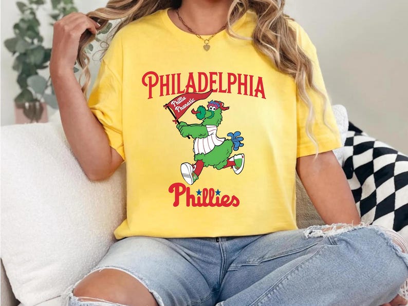 Może przedstawiać: Ż&oacute;łta koszulka z maskotką Philadelphia Phillies, zielonym, włochatym stworzeniem trzymającym flagę z napisem "Phillie Phanatic". Koszulka wyświetla r&oacute;wnież "Philadelphia" i "Phillies" w czerwonych literach.